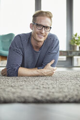 Porträt eines lächelnden jungen Mannes, der zu Hause auf einem Teppich liegt - RBF06849
