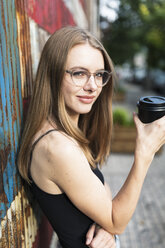 Junge Frau hält eine Tasse Kaffee in der Hand und steht vor einem Metallzaun mit Sternen und Streifen - GIOF04501