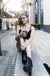 Junge Frau, die auf einer Mauer sitzt und einen Kaffee zum Mitnehmen trinkt - GIOF04504