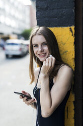 Young woman leaning on street corner, listening music, using smartphone - GIOF04517