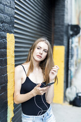 Young woman leaning on street corner, listening music, using smartphone - GIOF04518