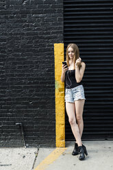 Junge Frau lehnt an einer Straßenecke, hört Musik und benutzt ein Smartphone - GIOF04519