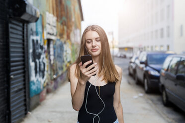 Young woman walking in street, listening music, using smartphone - GIOF04522