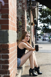 Young woman sitting in the street on a doorstep, holding a cup of coffee - GIOF04523