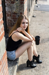 Young woman sitting in the street on a doorstep, holding a cup of coffee - GIOF04524