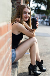 Young woman sitting in the street on a doorstep, holding a cup of coffee - GIOF04525