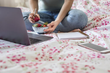 Frau sitzt auf dem Bett und isst Erdbeeren, während sie mit dem Laptop arbeitet, Teilansicht - FLMF00069