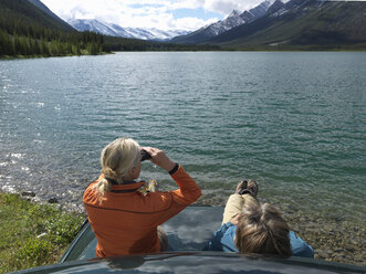 Ein Paar sitzt auf der Motorhaube eines Lastwagens und benutzt ein Fernglas über dem See - AURF06142