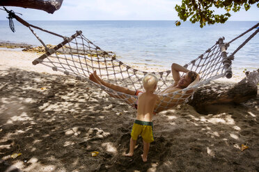 Vater in Hängematte und Sohn am Strand stehend, Bali, Indonesien - AURF06268