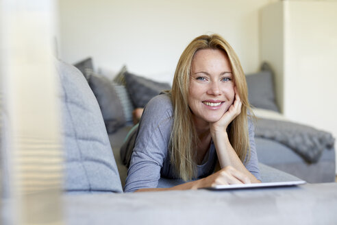 Lachende junge Frau, die zu Hause auf dem Sofa liegt und ein digitales Tablet benutzt - PNEF00947