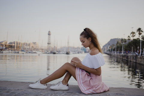 Modische junge Frau, die am Hafen von Barcelona sitzt und ihr Smartphone benutzt, lizenzfreies Stockfoto