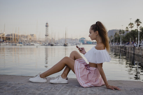 Modische junge Frau, die am Hafen von Barcelona sitzt und ihr Smartphone benutzt - AFVF01609