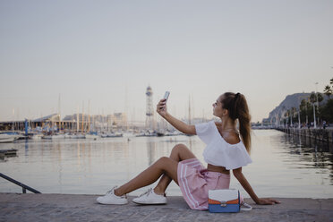 Modische junge Frau, die am Hafen von Barcelona sitzt und ihr Smartphone benutzt - AFVF01610