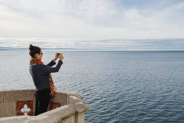 Seitenansicht einer jungen Frau, die das Meer mit ihrem Smartphone fotografiert, während sie auf einer Gebäudeterrasse vor dem Himmel steht - CAVF48771