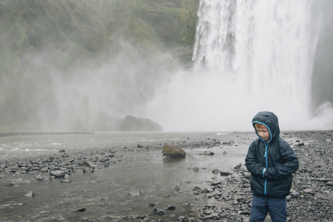 Boy wearing raincoat standing against splashing waterfall stock photo