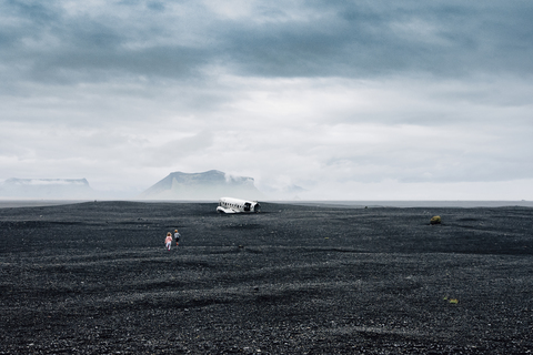 Mid distance view of siblings running towards wrecked airplane on black sand at beach against cloudy sky stock photo