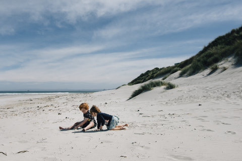 Full length of siblings playing with sand at beach against cloudy sky during sunny day stock photo