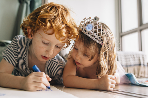 Brother drawing while lying by sister on floor at home stock photo