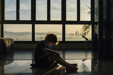 Full length of boy reading book while sitting on floor against windows at home stock photo