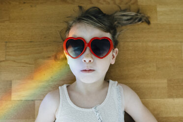 Draufsicht auf ein Mädchen, das eine herzförmige Sonnenbrille trägt und zu Hause auf dem Parkettboden liegt - CAVF48814