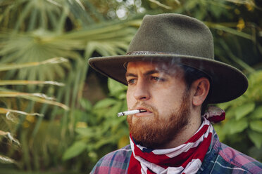 Close-up of bearded young man wearing hat and bandana while smoking cigarette - CAVF48838