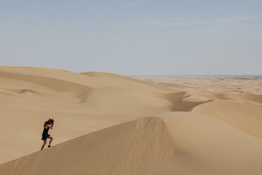 Seitenansicht einer jungen Frau mit Hut beim Klettern auf einer Sanddüne in der Wüste gegen den Himmel - CAVF48839