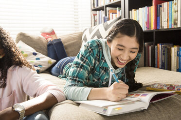 Fröhliches Teenager-Mädchen macht Hausaufgaben, während sie bei einem Freund zu Hause auf der Couch liegt - CAVF48853