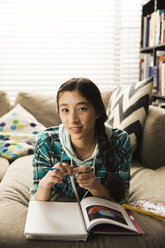 Porträt eines jungen Mädchens, das zu Hause auf der Couch liegend Hausaufgaben macht - CAVF48857