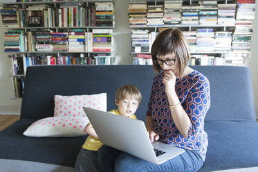 Mutter, die einen Laptop benutzt, während sie mit ihrem süßen Sohn zu Hause auf dem Sofa sitzt - CAVF48864