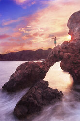 Rear view of shirtless man with arms outstretched standing on rock over sea against sky during sunset - CAVF48894