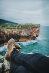 Low section of man sitting on mountain by sea against cloudy sky - CAVF48897