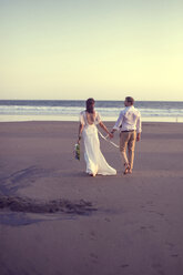 Rear view of newlywed couple holding hands while walking at beach against clear sky during sunset - CAVF48900