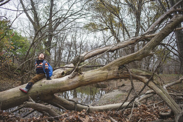 Boy with backpack sitting on fallen tree against sky in forest - CAVF48928
