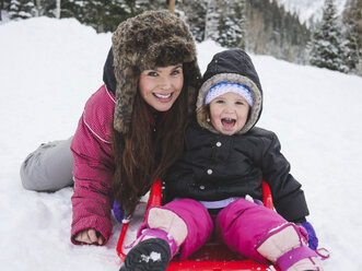 Portrait of happy mother with daughter sitting on snow covered field - CAVF48935