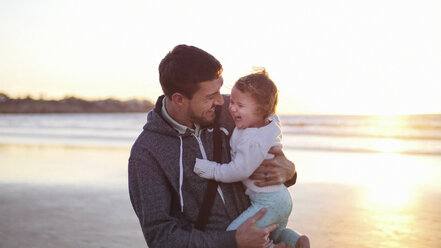 Glücklicher Vater, der seine Tochter trägt, während er am Strand gegen den klaren Himmel bei Sonnenuntergang steht - CAVF48937