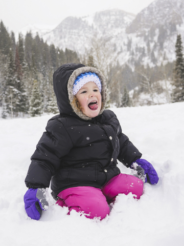 Glückliches kleines Mädchen kniend auf schneebedecktem Feld vor einem Berg, lizenzfreies Stockfoto