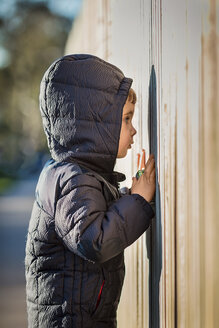 Seitenansicht eines neugierigen Jungen mit Kapuzenjacke, der durch einen Holzzaun späht - CAVF48951