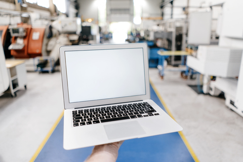 Hand hält Laptop mit leerem Bildschirm in einer Fabrikhalle, lizenzfreies Stockfoto