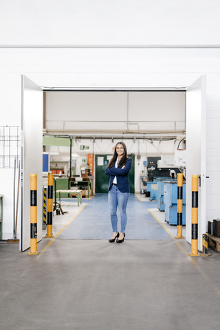 Selbstbewusste Frau, die in einem High-Tech-Unternehmen arbeitet, steht mit verschränkten Armen in einer Fabrikhalle, lizenzfreies Stockfoto