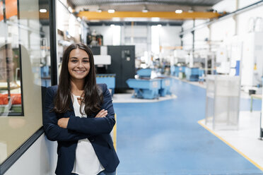 Selbstbewusste Frau, die in einem High-Tech-Unternehmen arbeitet, steht mit verschränkten Armen in einer Fabrikhalle - KNSF04968