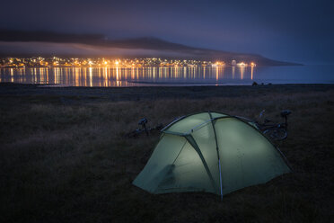 Zelt auf grasbewachsenem Strand, von innen beleuchtet, am Ufer des Ozeans und der Lichter der Stadt im Hintergrund - AURF06480