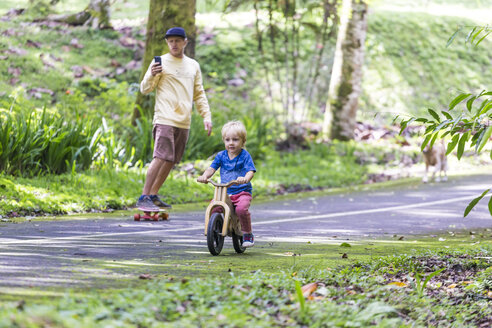 Father on skateboard with phone accompanying son on bicycle - AURF06870