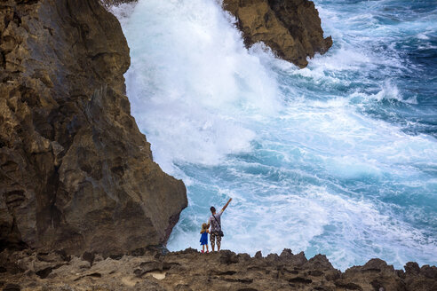 Vater und Tochter stehen auf Küstenklippen und schauen auf plätschernde Wellen, Bali, Indonesien - AURF06875