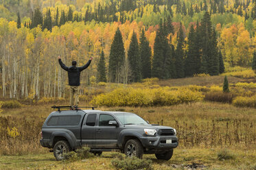 Mann steht auf dem Dach eines Geländewagens mit erhobenen Armen vor einem Wald mit Herbstfarben, Crested Butte, Colorado, USA - AURF07095