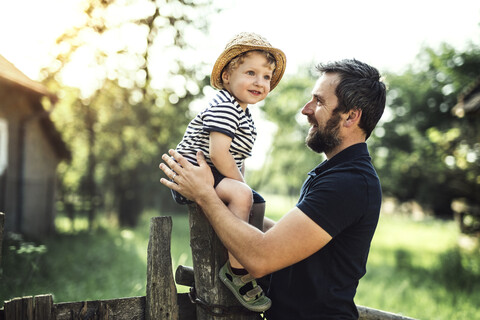 Glücklicher Vater betrachtet seinen lächelnden kleinen Sohn, der auf einem Zaun sitzt, lizenzfreies Stockfoto