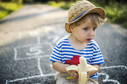 Porträt eines kleinen Jungen mit Banane, der auf der Straße sitzt und etwas beobachtet, lizenzfreies Stockfoto