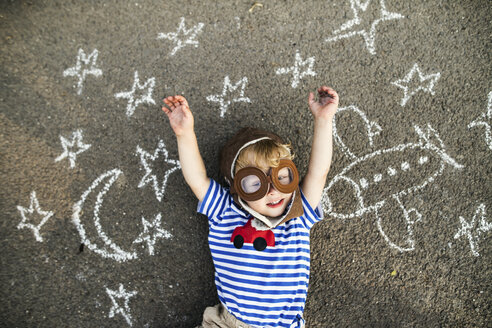 Portrait of smiling toddler wearing pilot hat and goggles lying on asphalt painted with airplane, moon and stars - HAPF02779