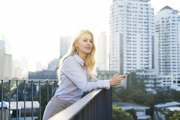 Blonde smiling business woman leaning onto handrail holding smartphone on city rooftop - SBOF01506