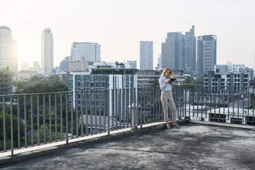 Blonde Geschäftsfrau lehnt sich an ein Geländer und hält ein Tablet auf dem Dach der Stadt - SBOF01510