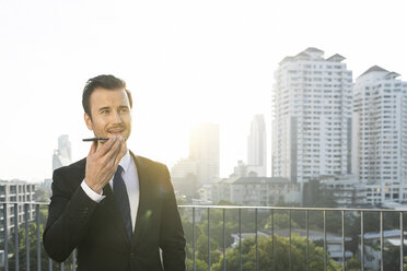 Business man in dark suit speaking into smartphone on city rooftop - SBOF01519